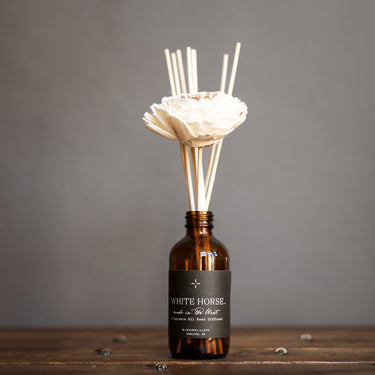 A White Horse reed diffuser with a bottle of scented diffuser oil and several reeds inserted in sola wood flowers, arranged on a wooden surface against a grey background.