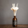 A White Horse reed diffuser with a bottle of scented diffuser oil and several reeds inserted in sola wood flowers, arranged on a wooden surface against a grey background.