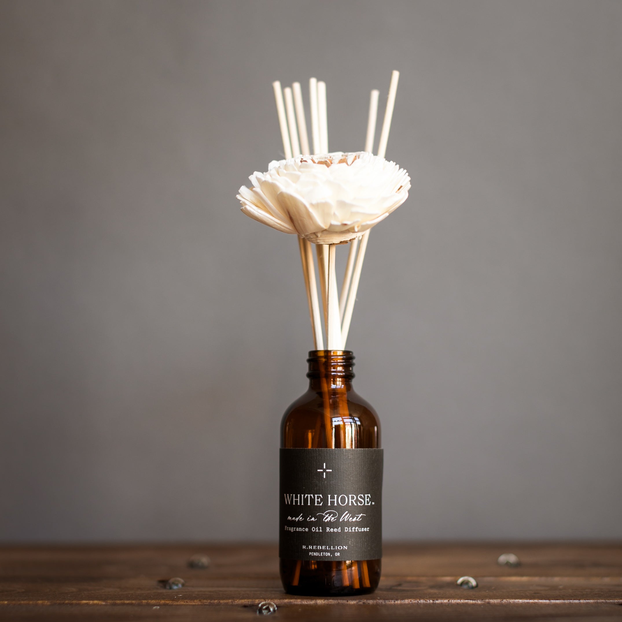 A White Horse reed diffuser with a bottle of scented diffuser oil and several reeds inserted in sola wood flowers, arranged on a wooden surface against a grey background.